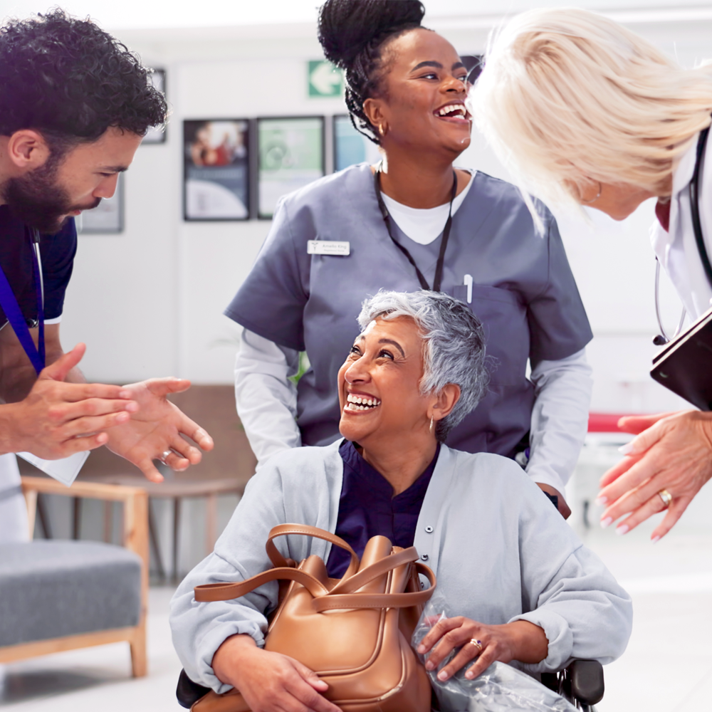 A patient is smiling as they leave a care facility with nurses, aides, and a physician in tow.