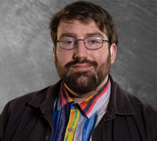A man smiles at the camera with a beard, glasses, and short brown hair. He has a watercolor rainbow button up shirt, and a black denim jacket over it.