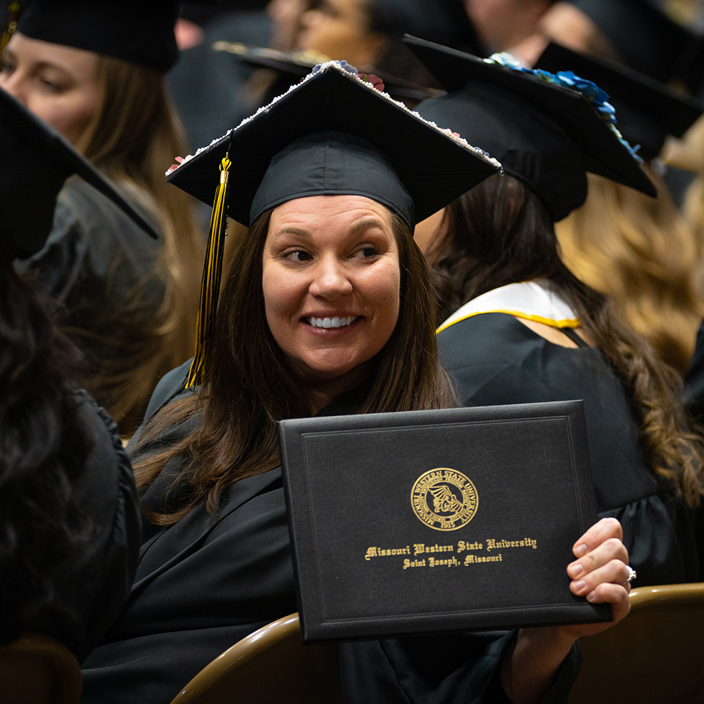 graduate smiling and holding diploma cover while seated at commencement