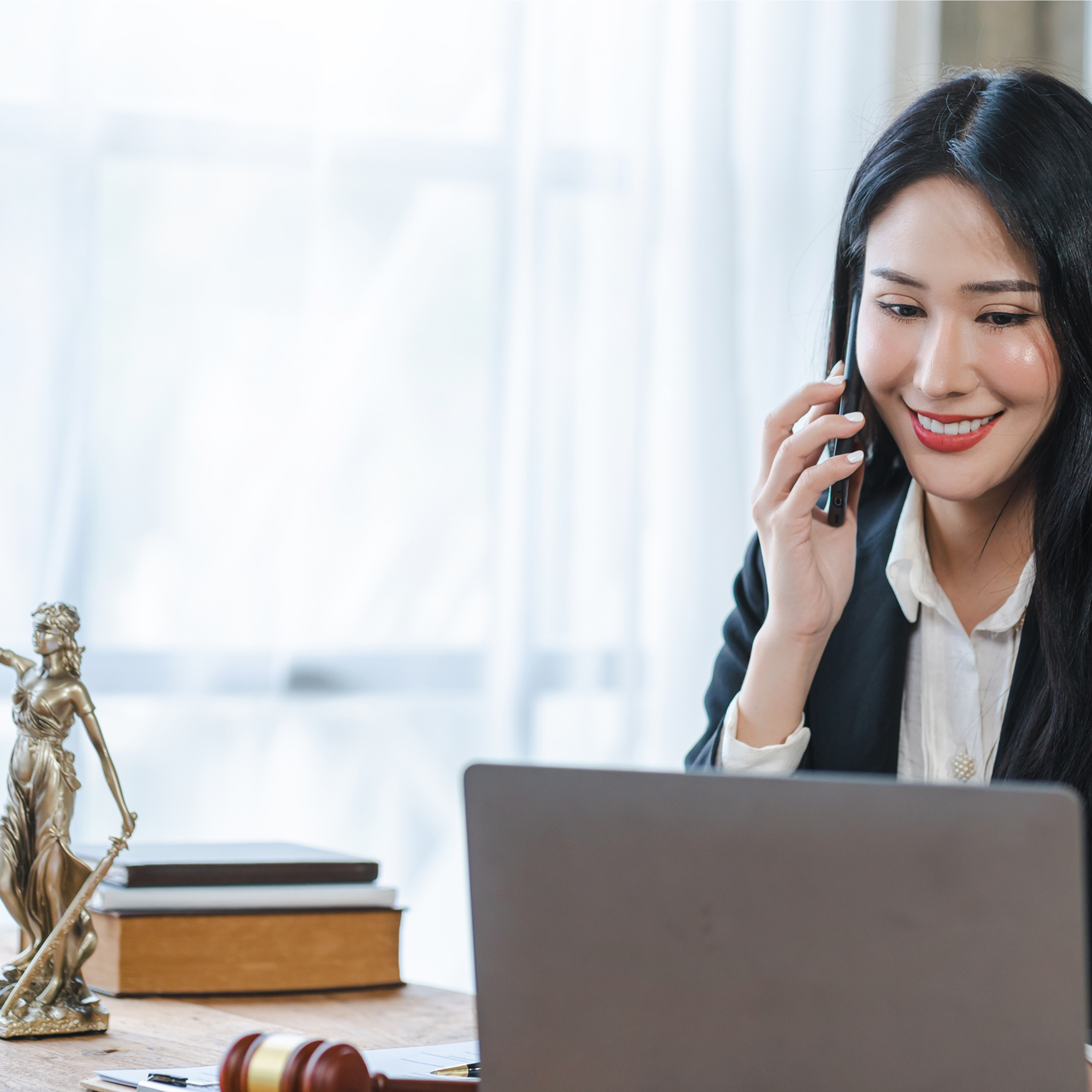 Paralegal Certificate 1 A woman sits at her laptop speaking on the phone and smiling, a statue of lady justice beside her.