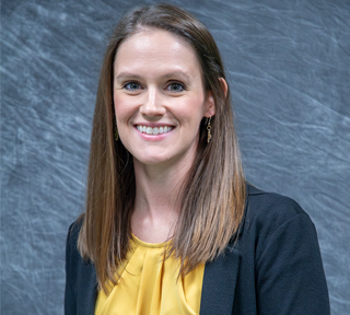 A woman smiles at the camera wearing a gold shirt and a black sweater with long brown hair.