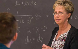 professor instructing a class with math equations on a chalk board in the background