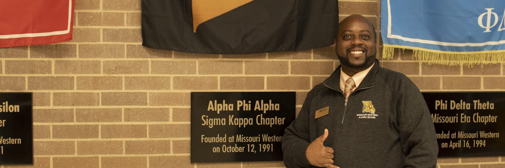 Lai-Monté Hunter posing in front of the Alpha Phi Alpha Sigma Kappa Chapter flag and plaque