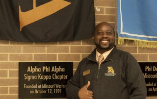 Lai-Monté Hunter posing in front of the Alpha Phi Alpha Sigma Kappa Chapter flag and plaque