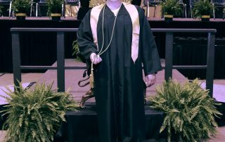 Allison Hildebrand posing for a photo in her cap and gown with commencement stage in background