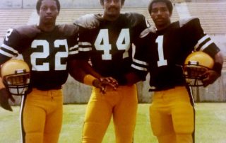 Tim Hoskins, Rodney Stephenson and Marc Lewis posing for photo in football uniforms, spratt stadium in background