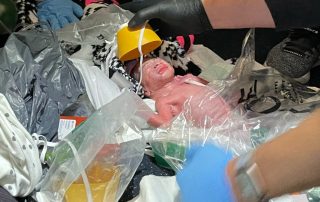 newborn baby surrounded by gloved hands, airplane oxygen mask, blankets and plastic bags