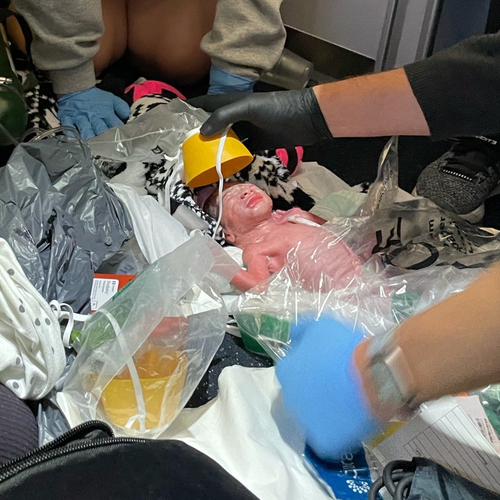 newborn baby surrounded by gloved hands, airplane oxygen mask, blankets and plastic bags