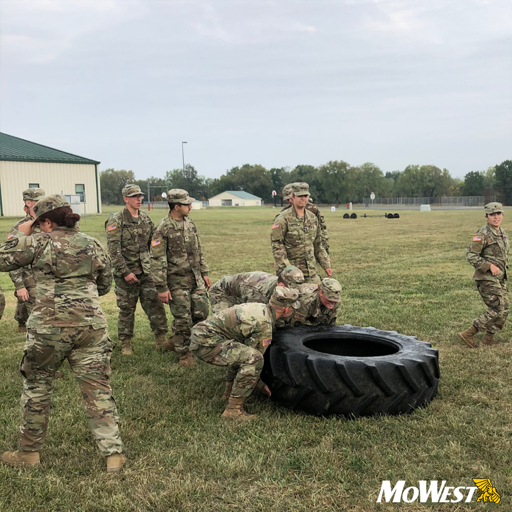 Students Flipping Tire Students in green camo fatigues work together to flip a tractor tire in a field on the University campus.
