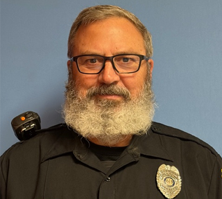 A police officer stands with a black shirt, a badge, and his radio. He has a white beard, sandy grey hair that is short, and glasses.