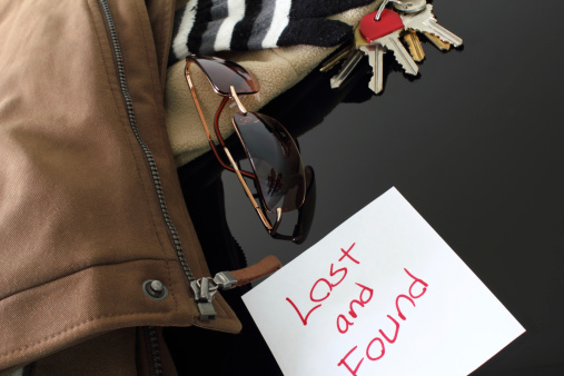 Multiple items (house keys, sunglasses, cotton stripped glove, coat) on a reflective table with a lost and found sign.