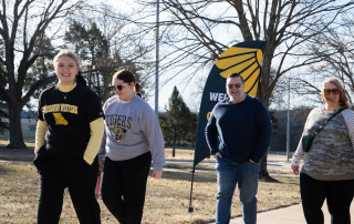 Four people walk past a Welcome Home banner on campus, smiling.