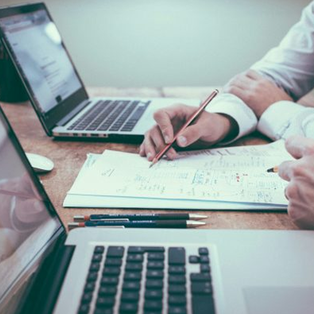 Two people sit with two laptops at a desk going over paperwork.