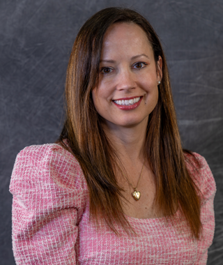 A woman with long brown hair and brown eyes smiles while wearing a pink dress and a gold locket.