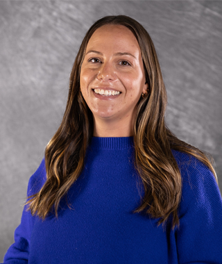 A woman with long brown hair and brown eyes smiles while wearing a blue knit sweater