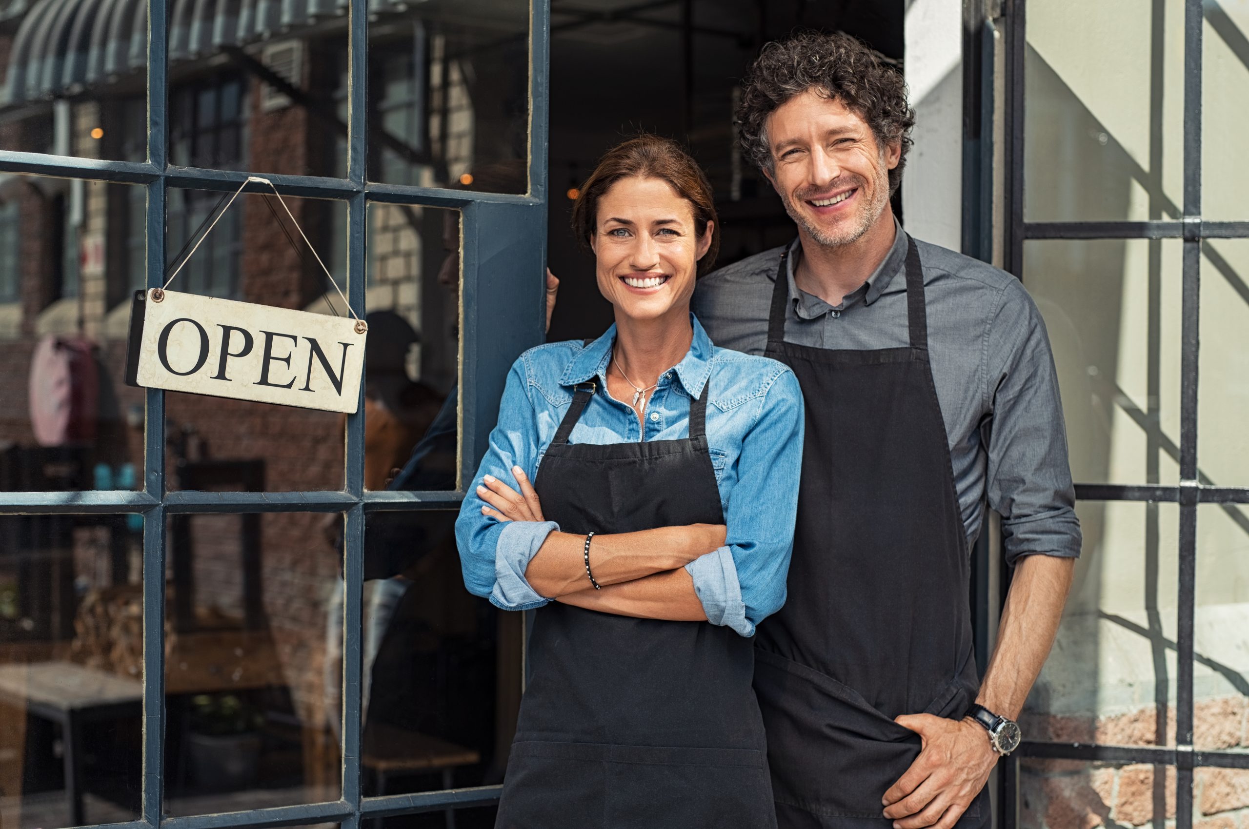 For Existing Businesses Two cheerful small business owners smiling and looking at camera while standing at entrance door. Happy mature man and mid woman at entrance of newly opened restaurant with open sign board. Smiling couple welcoming customers to small business shop.