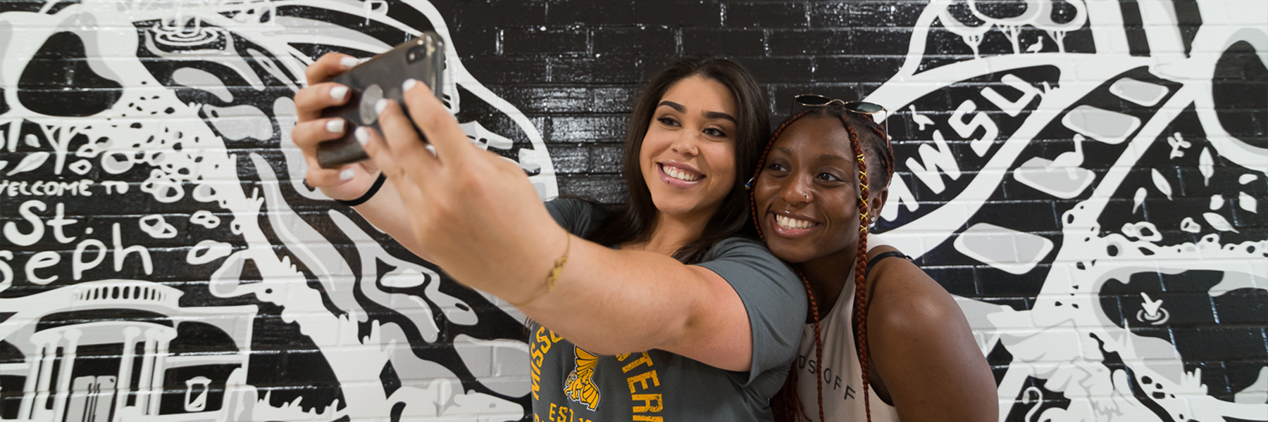 two students taking a selfie in front of a mural of wings