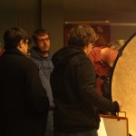 Students of Motion Picture Lighting gather around the lighting equipment as house lights get dimmer to observe contrast. Professor Hanson adjusts the settings on the tripod camera as a demonstration.